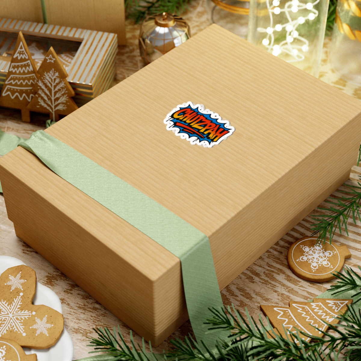 Gift box with a green ribbon and a colorful chutzpah sticker on a festive background with cookies and lights.