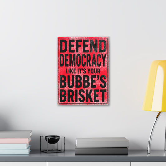 Framed poster with text 'Defend Democracy Like It's Your Bubbe's Brisket' on a wall above a desk.