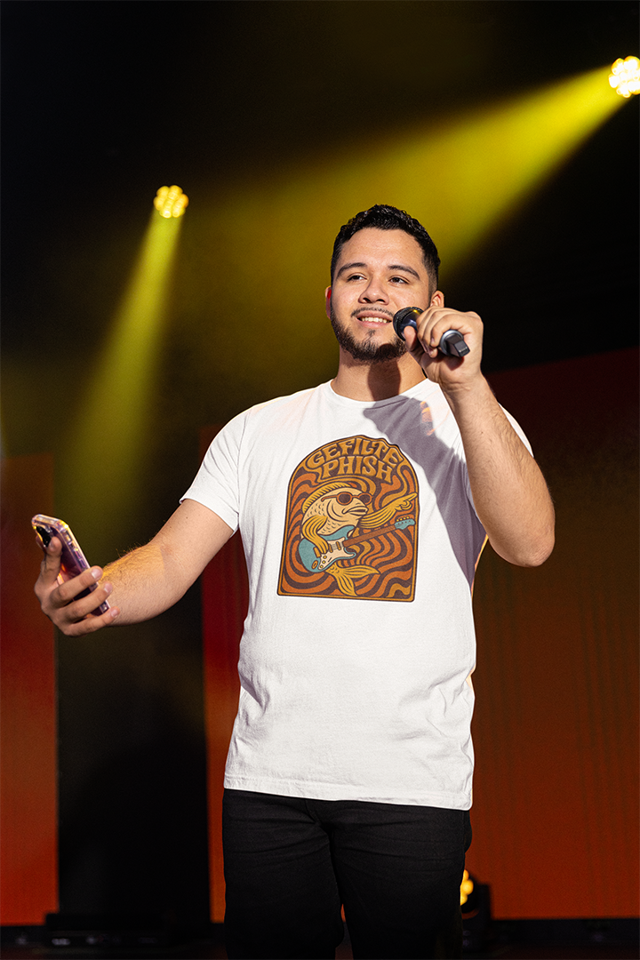 Man on stage holding a microphone and phone with a graphic t-shirt, illuminated by stage lights.