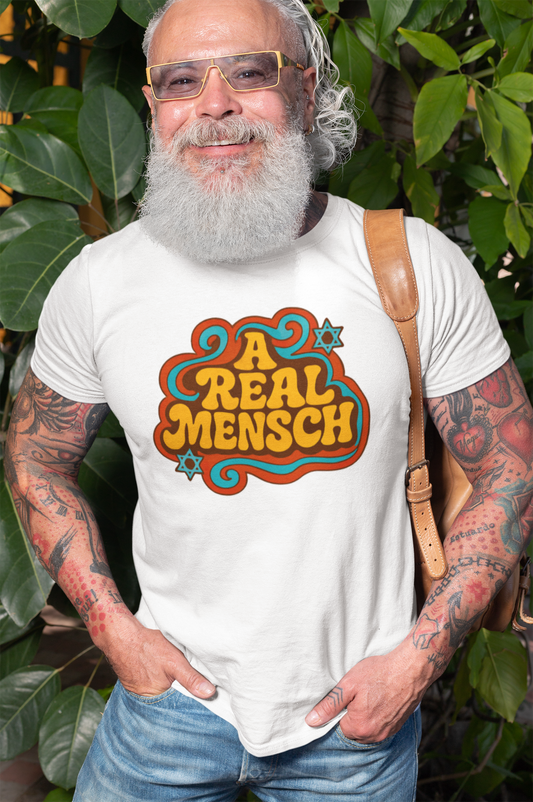 Man wearing a white t-shirt with 'A Real Mensch' design, standing outdoors with greenery.