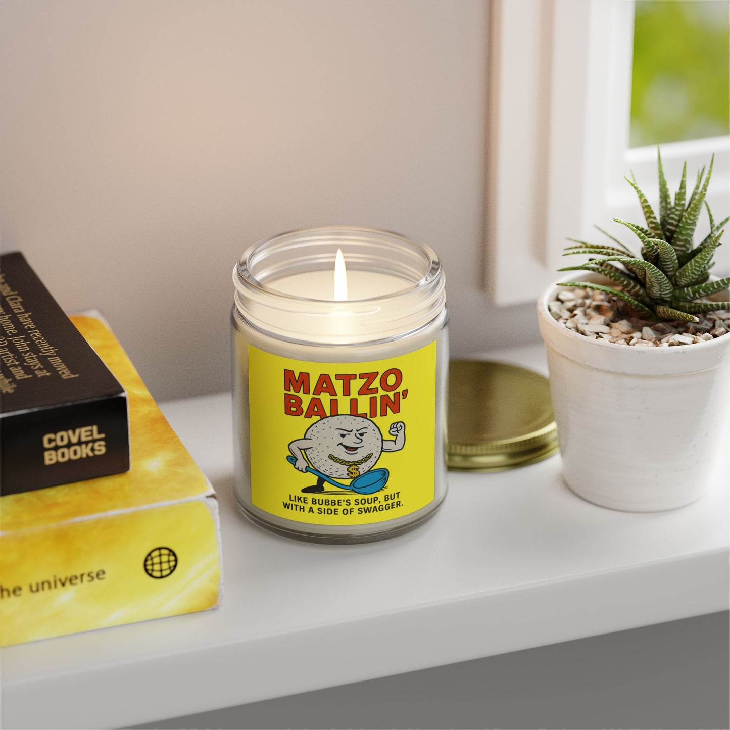 Candle with a 'Matzo Ballin' label on a shelf with books and a plant
