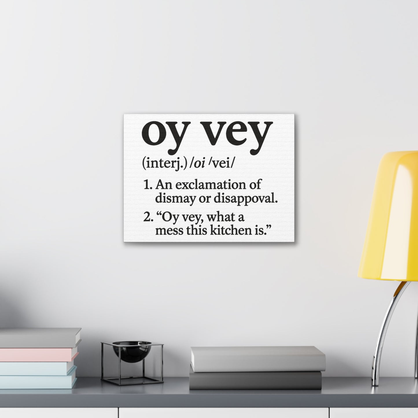 Canvas print with 'oy vey' definition on a wall above a desk with books and a lamp.