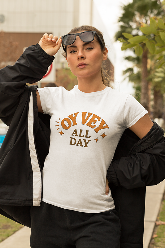 Person wearing a white t-shirt with 'Oy Vey All Day' text, holding sunglasses outdoors.