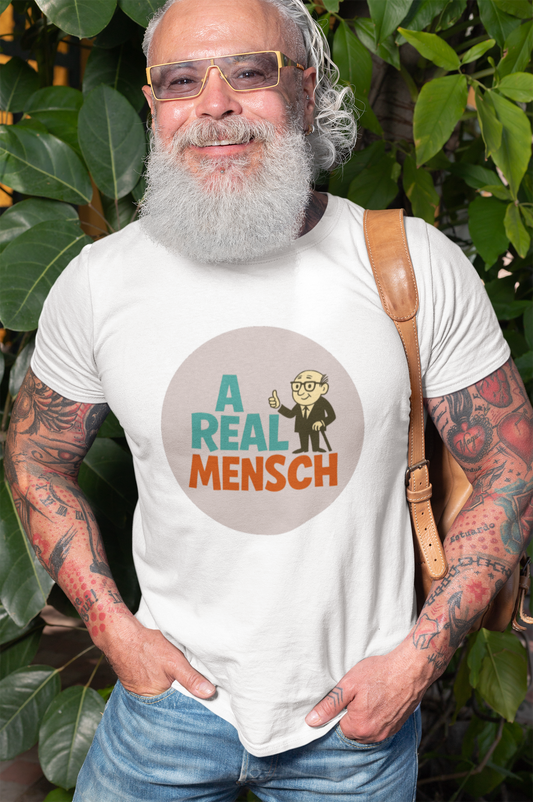 Man wearing a white t-shirt with a graphic and text 'A Real Mensch', standing outdoors with greenery in the background.