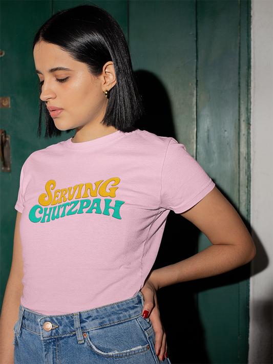 Woman wearing a pink t-shirt with 'Serving Chutzpah' text, standing against a green door.