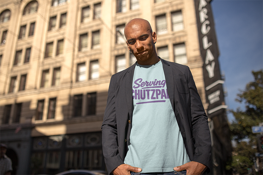 Man wearing a light blue t-shirt with purple 'serving chutzpah' text standing in front of a building.