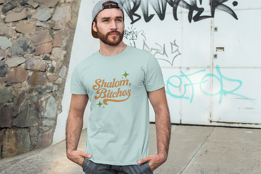 Man wearing a light green t-shirt with 'Shalom, Bitches' text in front of a graffiti-covered wall.