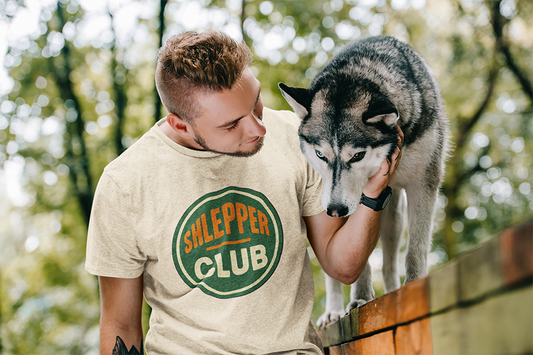 Man wearing a 'Shlepper Club' t-shirt petting a dog outdoors.
