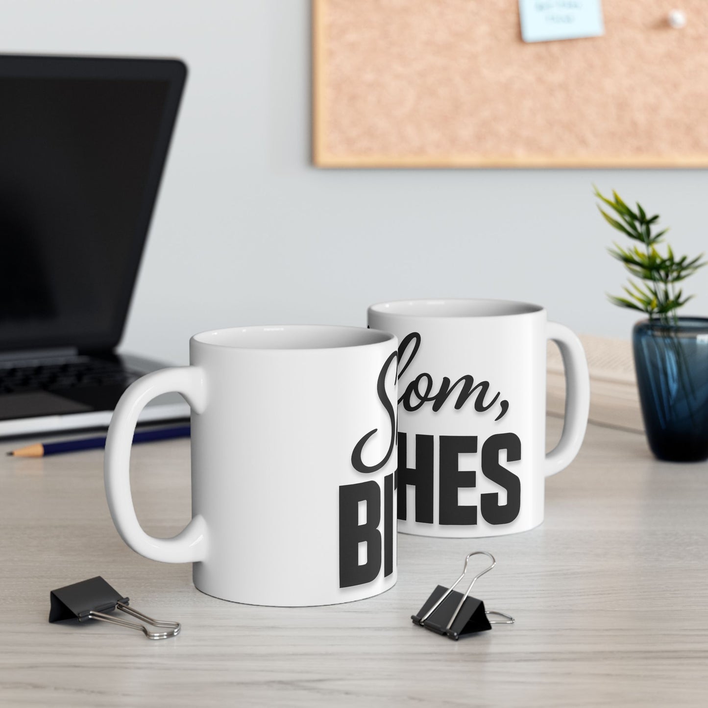 Two white mugs with black text 'shalom, bitches' on a desk with office supplies and a plant.
