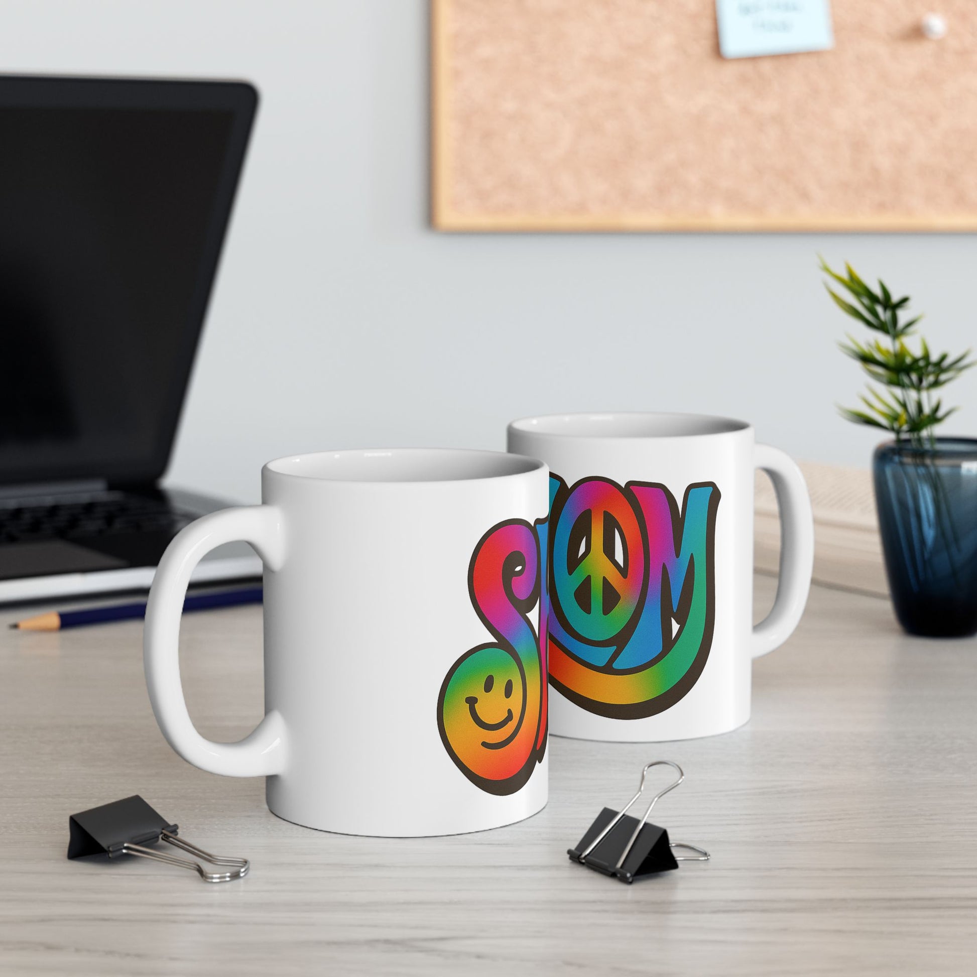 Two white mugs with colorful 'shalom' designs on a desk with a laptop and office supplies.