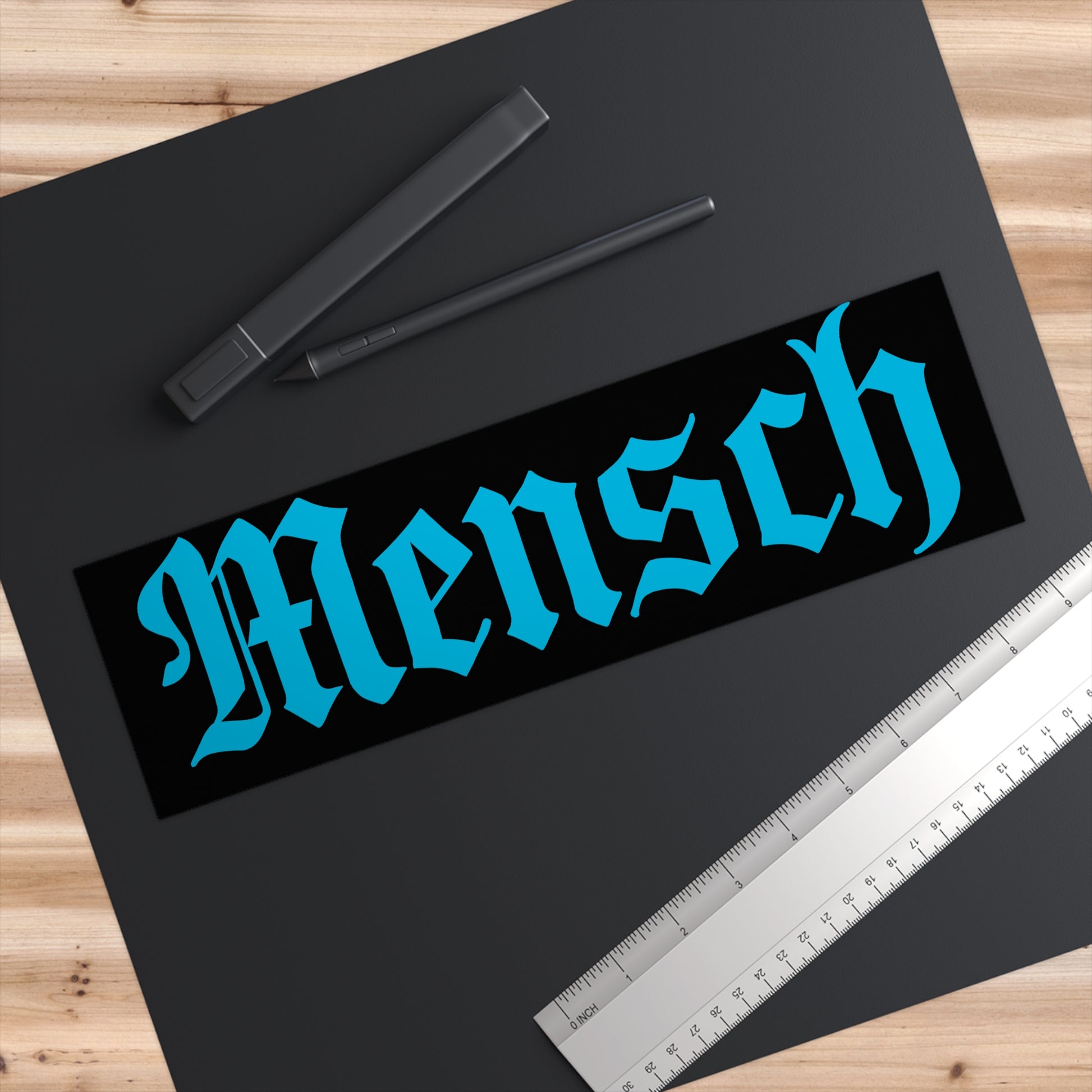 Black mat with 'Mensch' text in blue, pens, and a ruler on a wooden surface