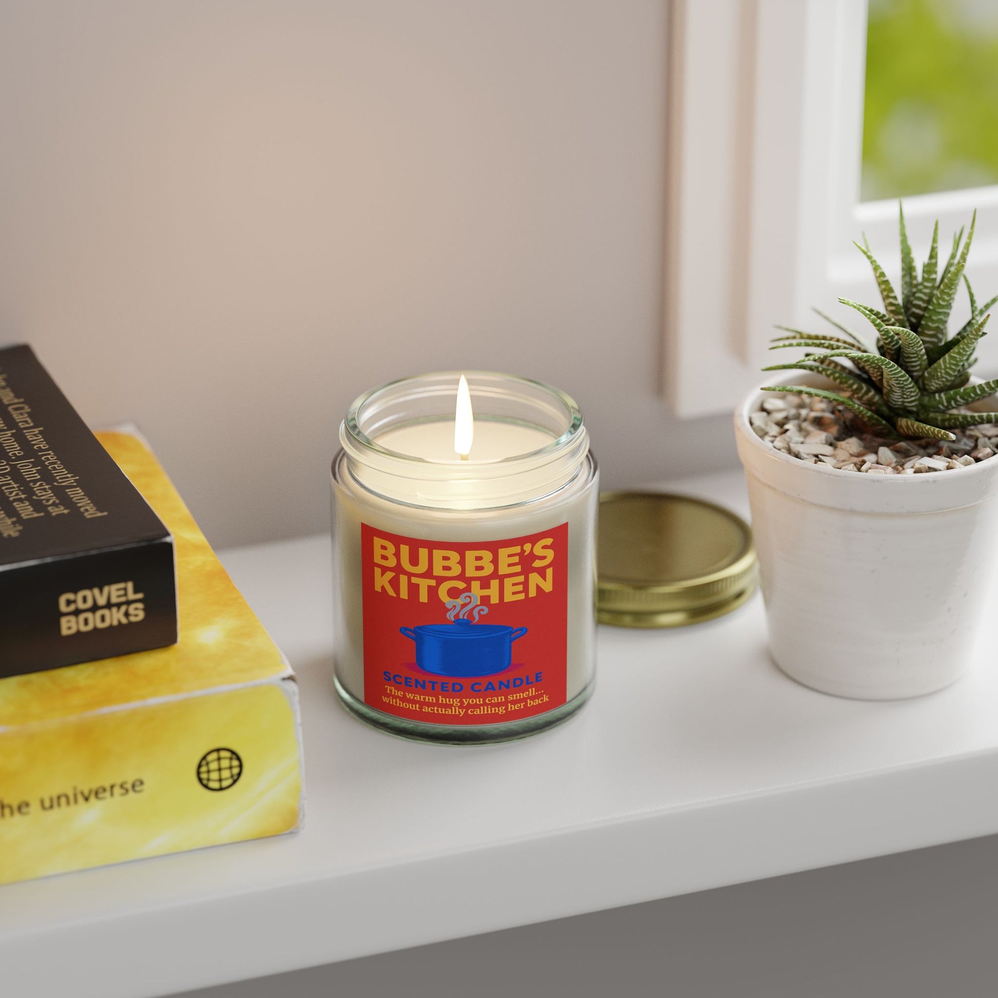 Candle labeled 'Bubbe's Kitchen' on a shelf with books and a plant