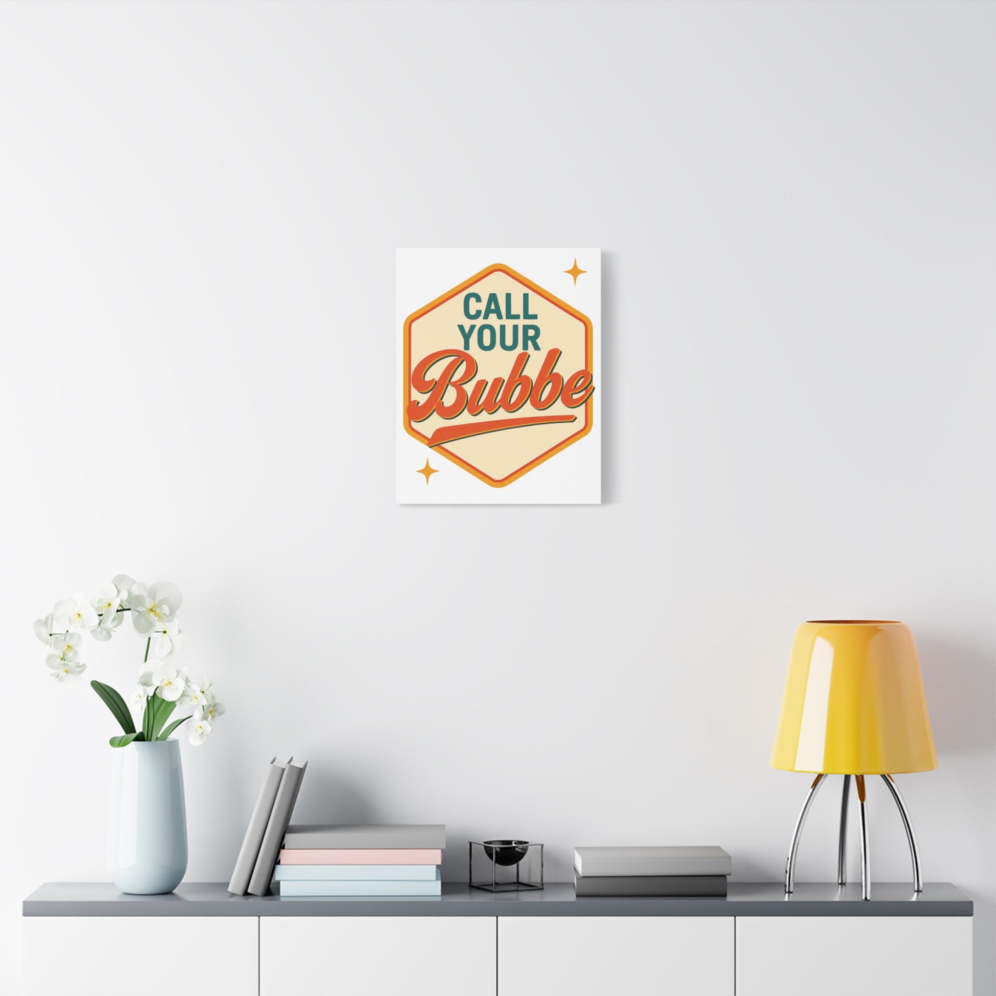 Decorative wall art with 'Call Your Bubbe' text on a white wall above a shelf with a lamp and books.