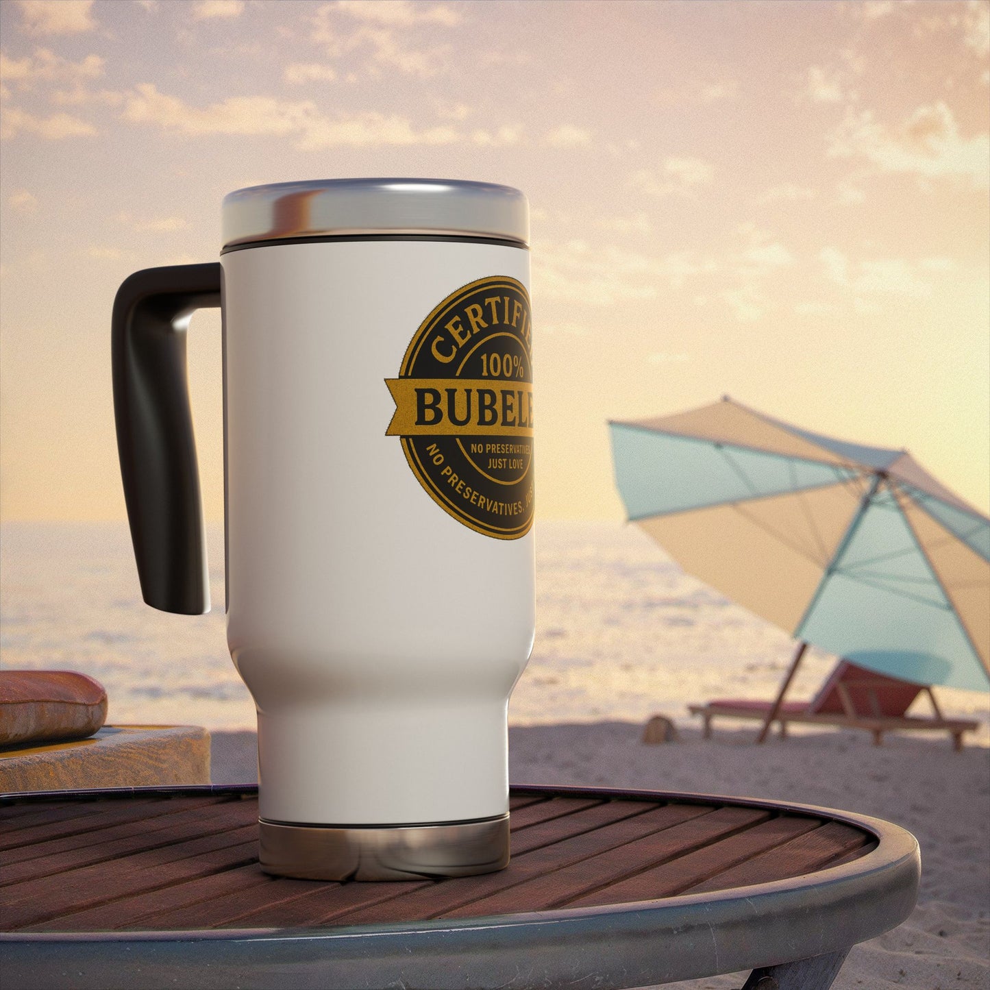 White travel mug with a gold 'certified bubeleh' label on a beach table with an umbrella and chairs in the background.