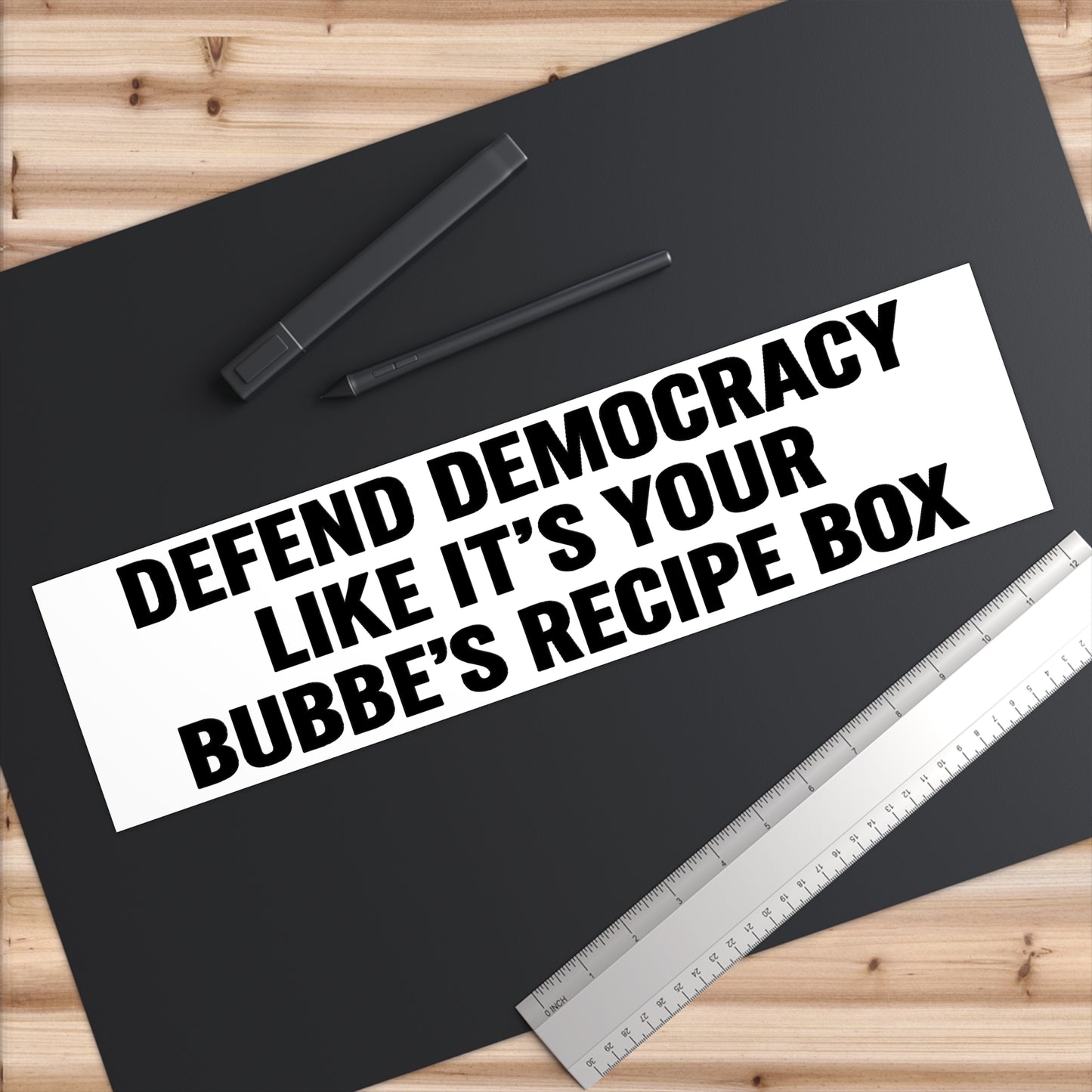 Black mat with text 'Defend democracy like it's your Bubbe's recipe box' on a wooden surface with a pen and ruler.