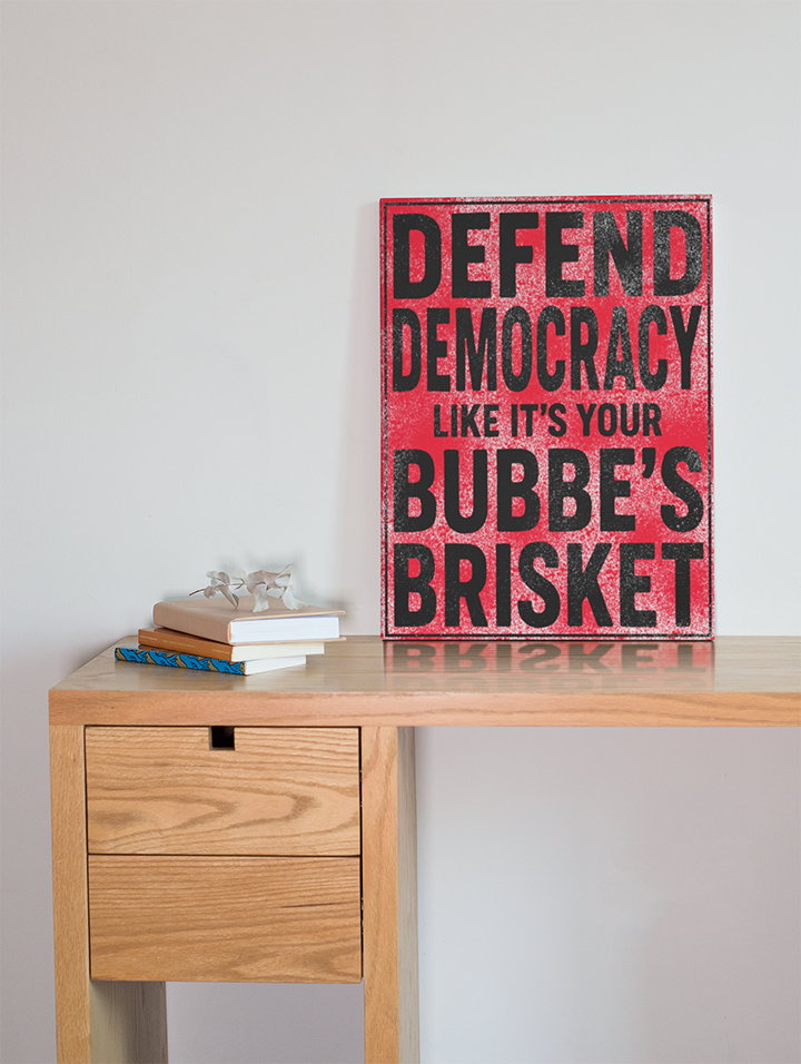 Wooden desk with a sign reading 'Defend Democracy like it's your Bubbe's Brisket' on a plain background