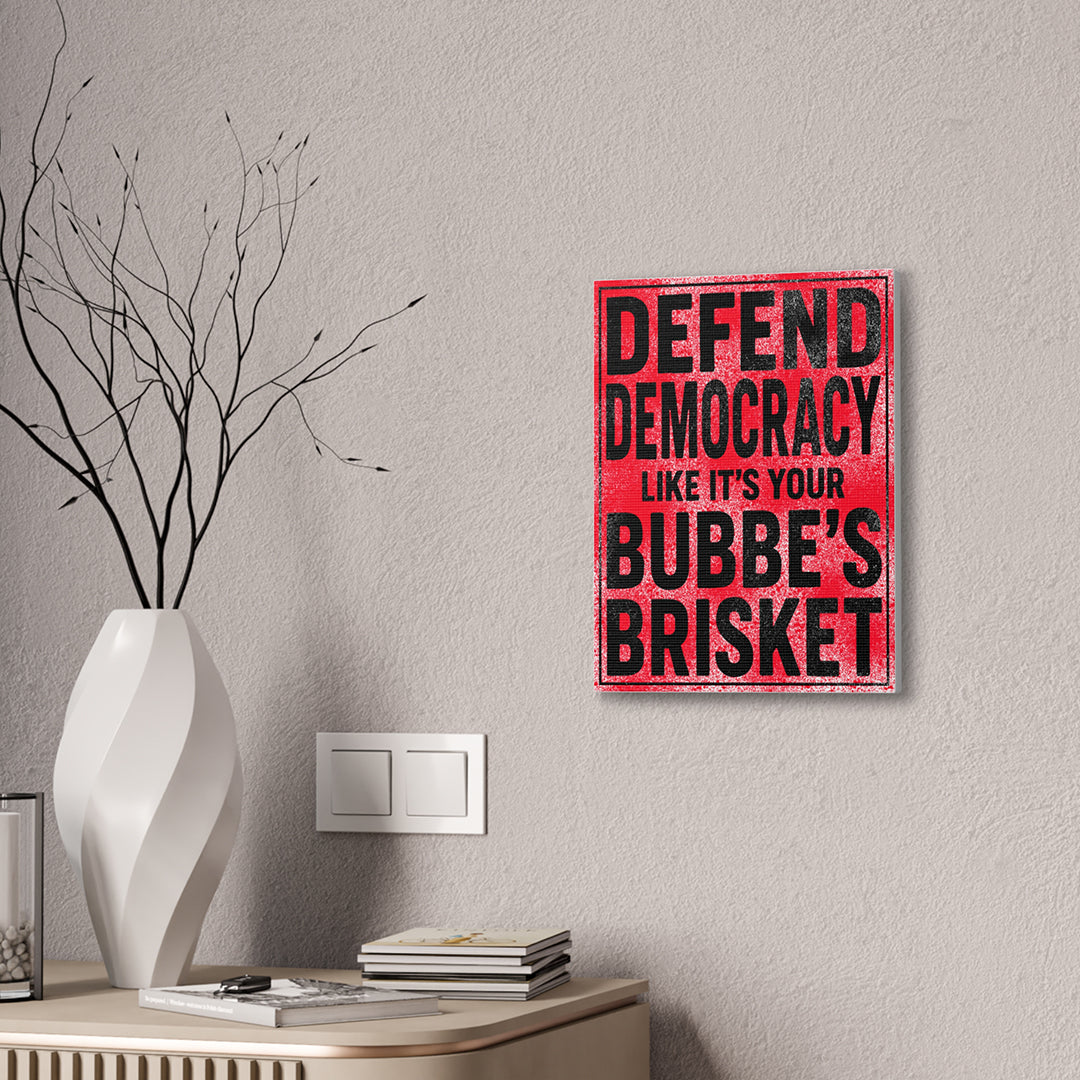 Wall art with text 'Defend Democracy like it's your Bubbe's Brisket' on a gray wall above a white vase and books.