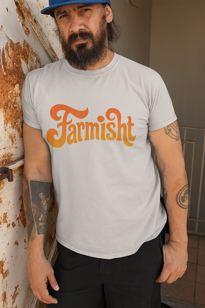Man wearing a gray t-shirt with 'Farmisht' printed in orange, leaning against a rusty wall.