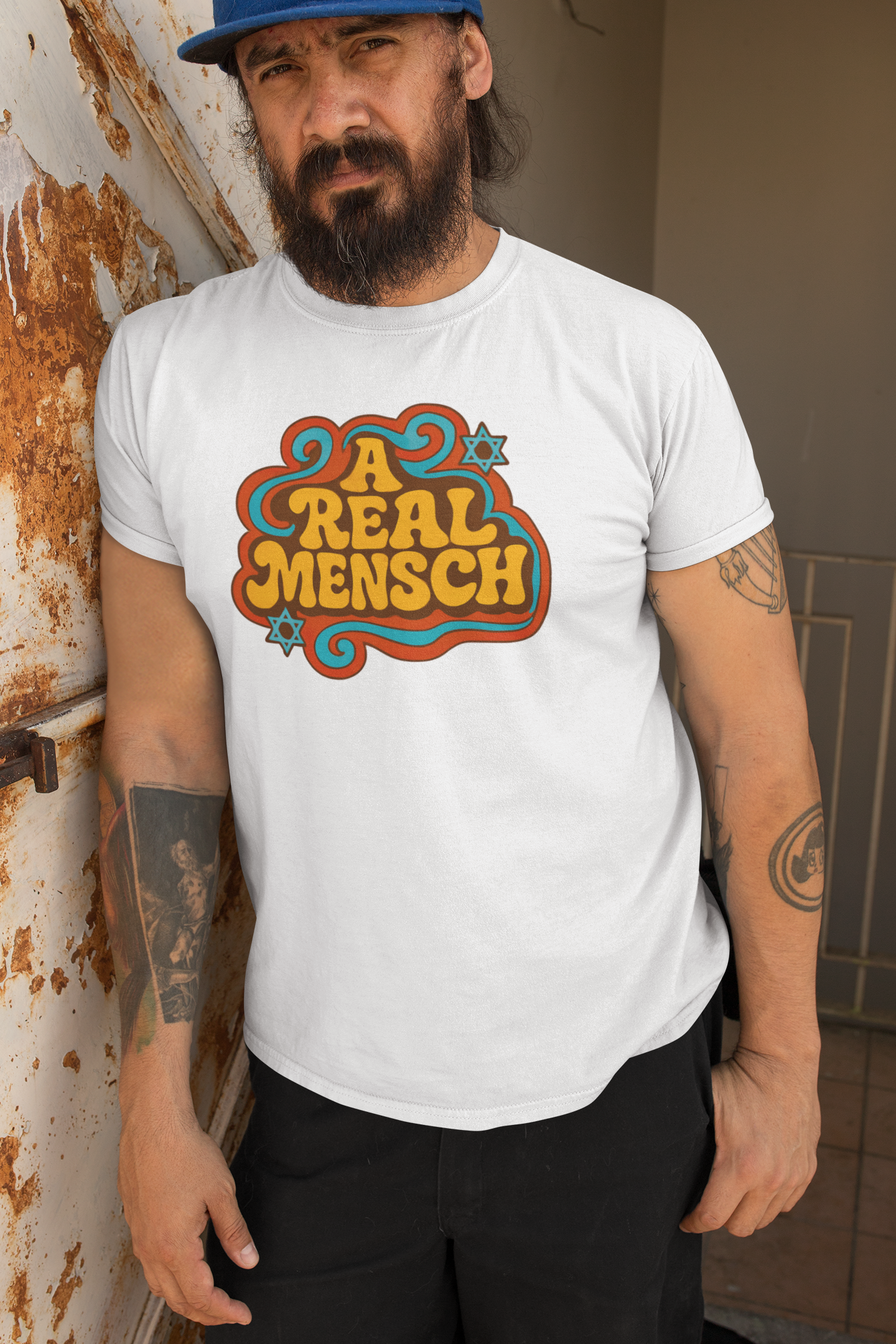 Man wearing a white t-shirt with 'A Real Mensch' design, standing against a rusty metal wall.