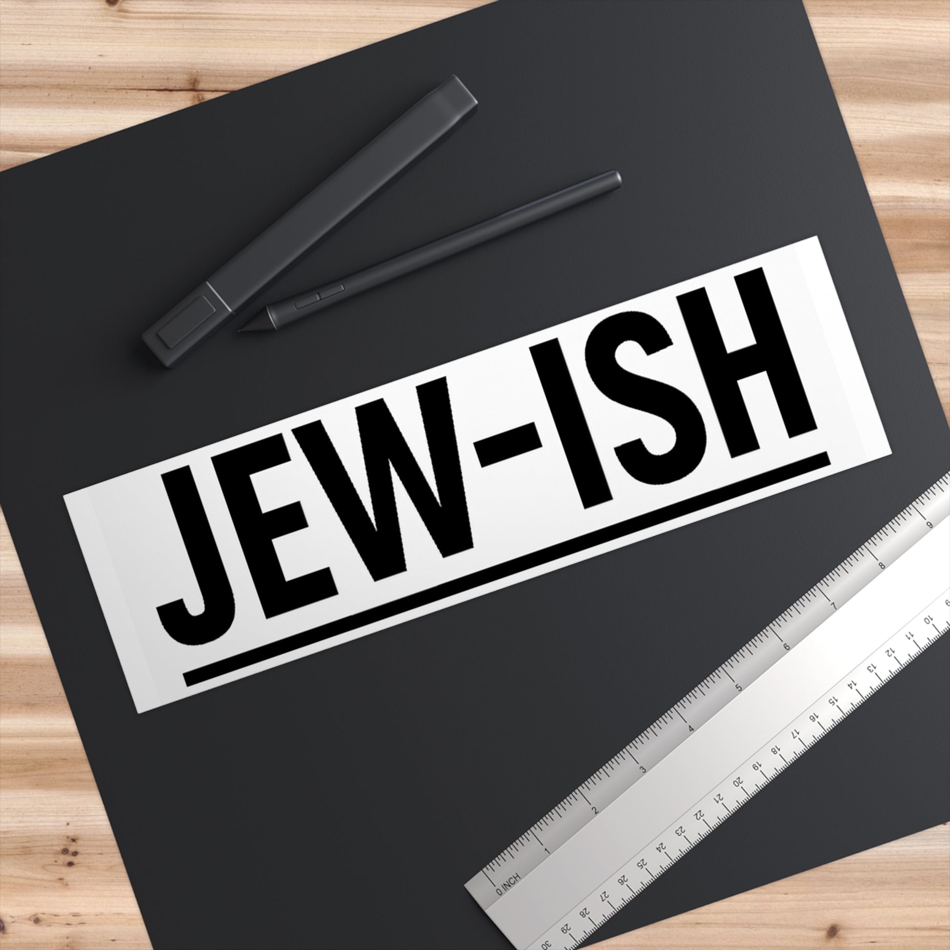 Sticker with 'JEW-ISH' text on a black surface with pens and a ruler.