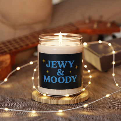 Candle in a jar with 'JEWY & MOODY' text on a wooden coaster surrounded by string lights.