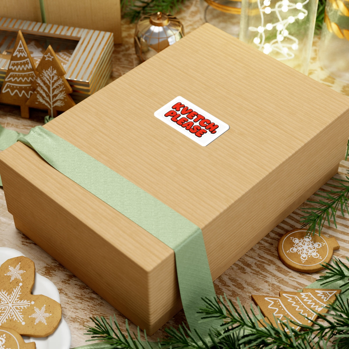 Gift box with a green ribbon and 'Kvetch please' label on a festive background with cookies and lights.