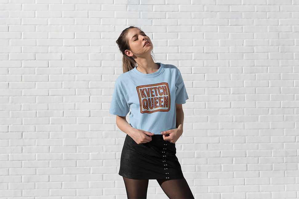 Woman wearing a light blue t-shirt with 'Kvetch Queen' text against a white brick wall.