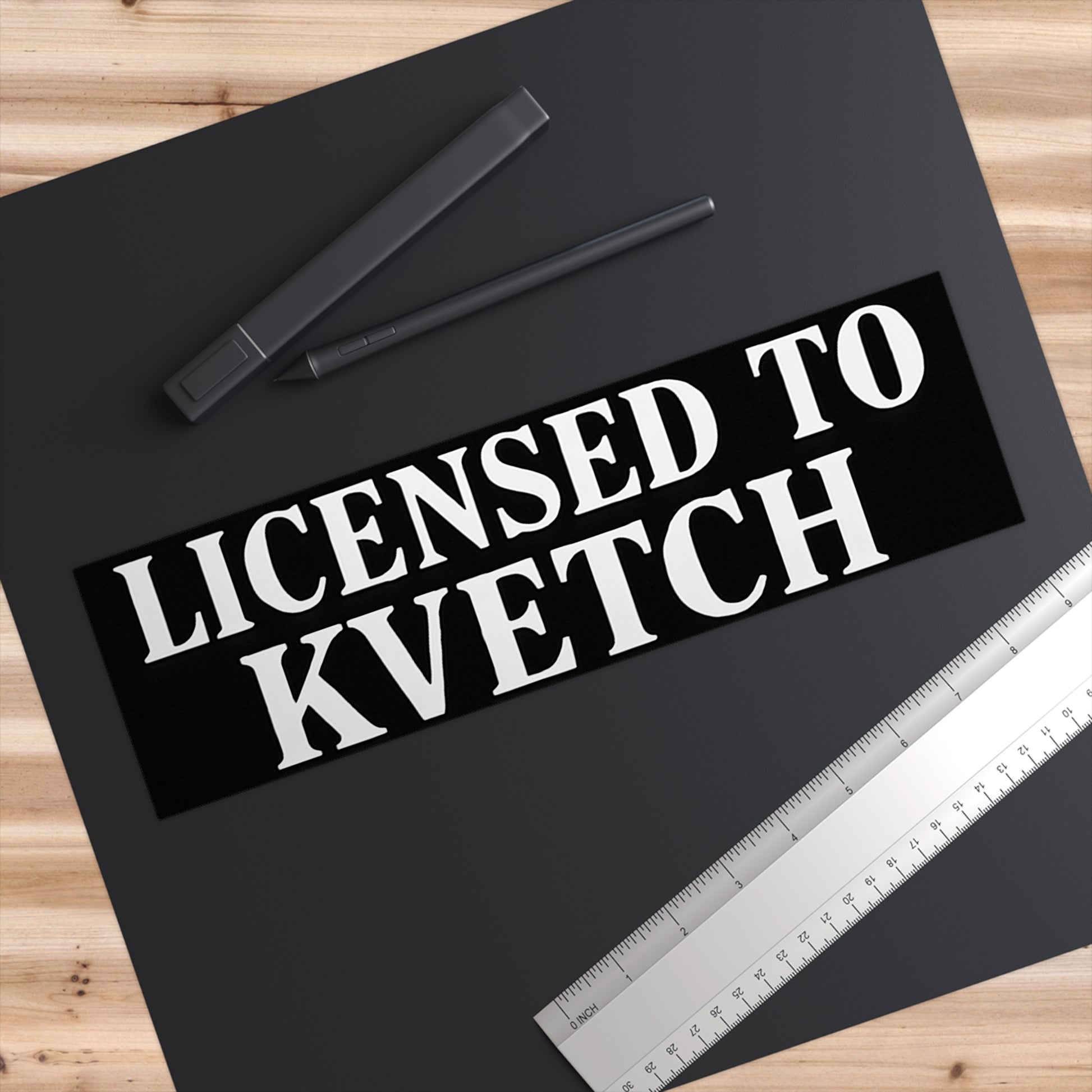 Black bumper sticker with 'LICENSED TO KVETCH' text on a black surface with pens and a ruler.