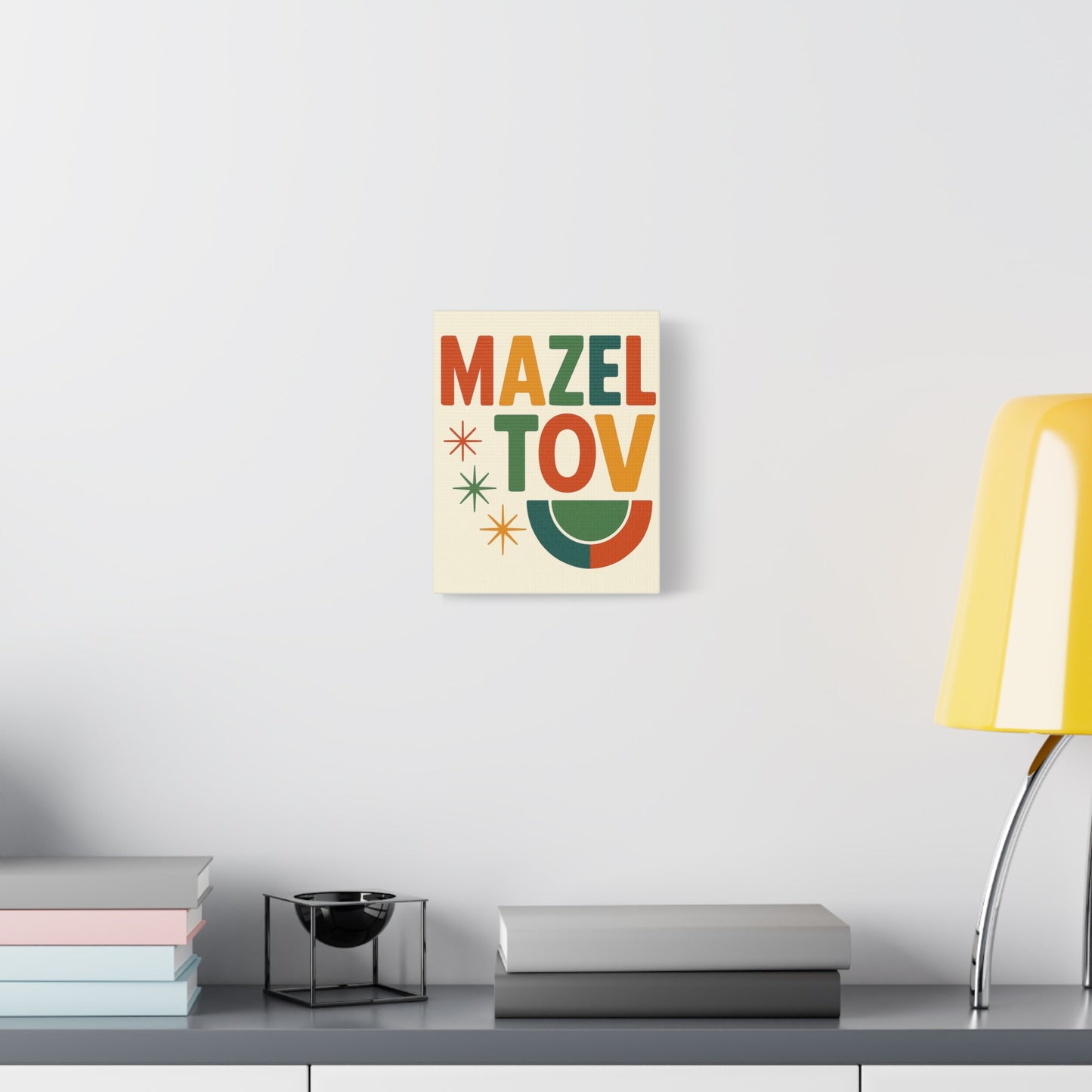Colorful 'Mazel Tov' print on a wall above a desk with books and a lamp.