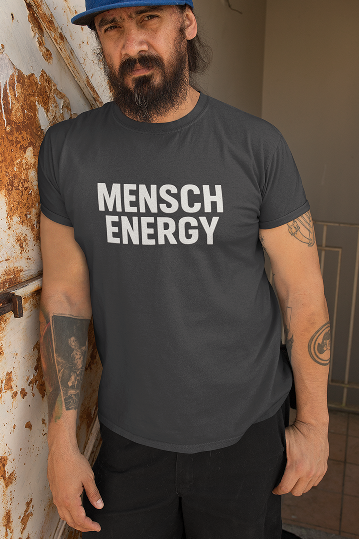Man wearing a gray 'MENSCH ENERGY' t-shirt leaning against a wall.