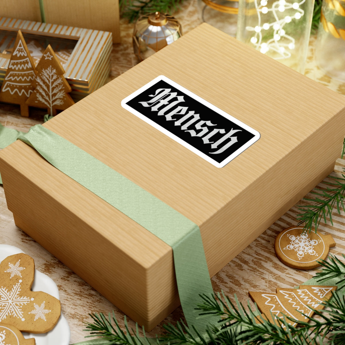 Wooden gift box with a green ribbon and 'Mensch' sticker on a festive background with cookies and lights.