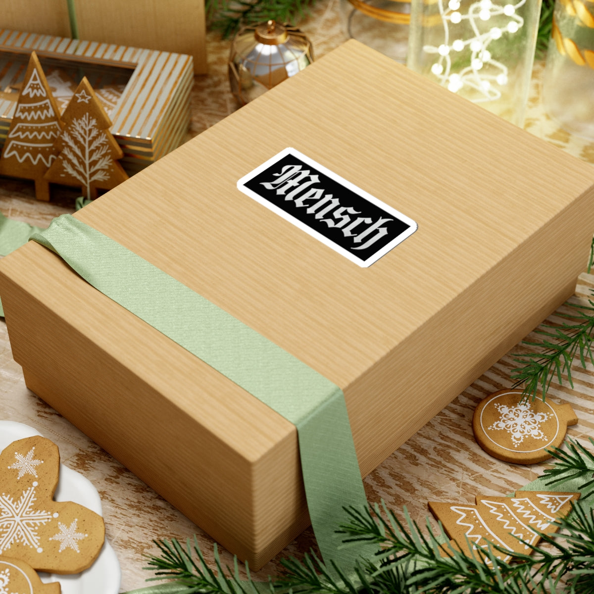 Wooden gift box with a green ribbon and a 'mensch' sticker on a festive background with cookies and decorations.
