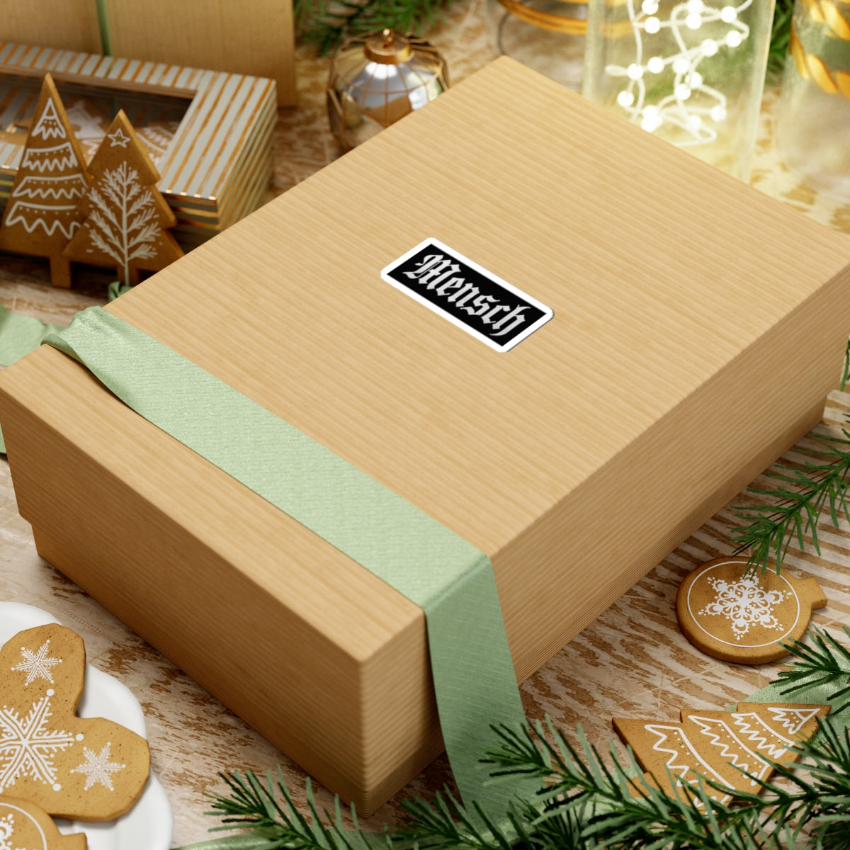 Gift box with a green ribbon and a 'mensch' sticker on a festive background with cookies and decorations.