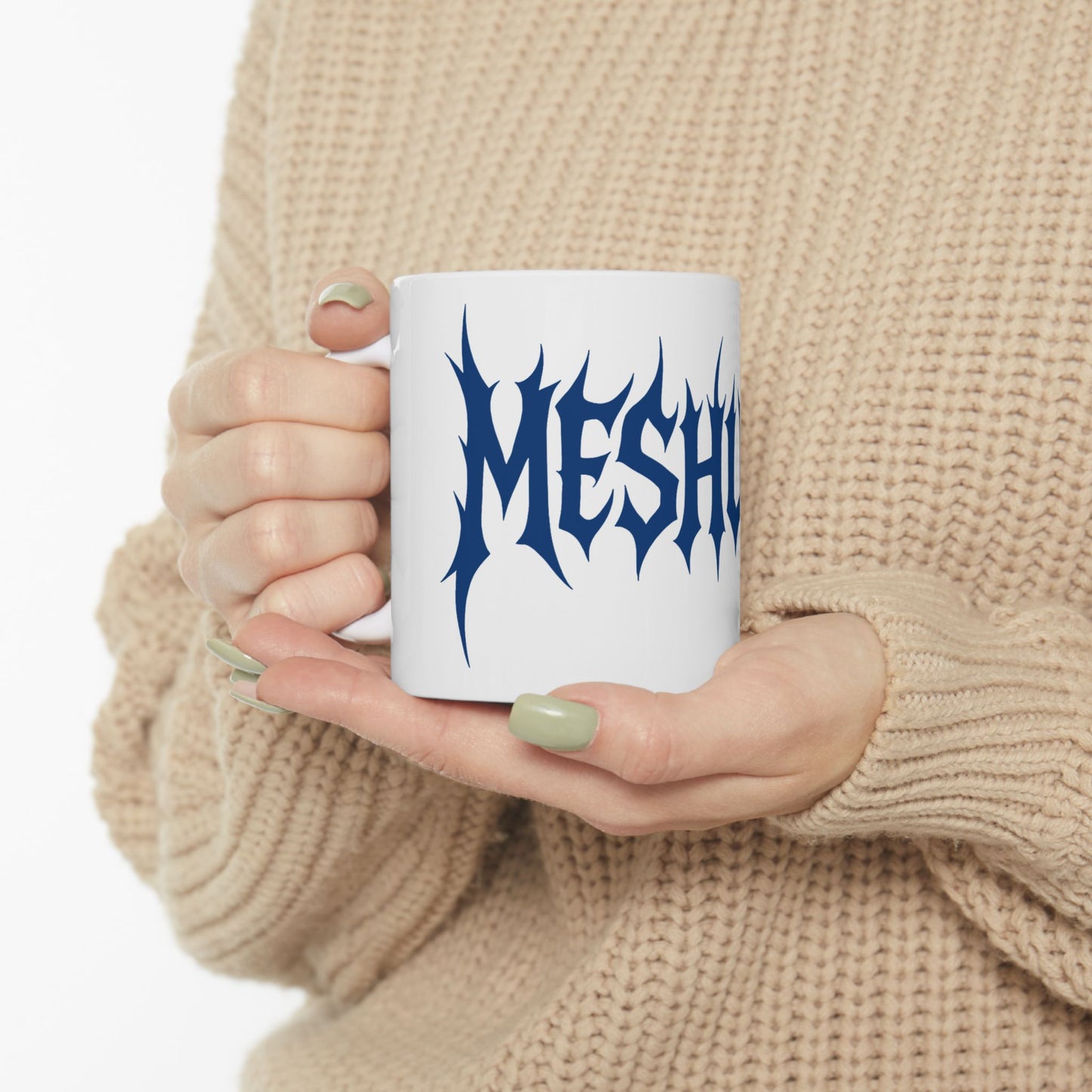 Person holding a white mug with blue text 'meshuga' against a beige background