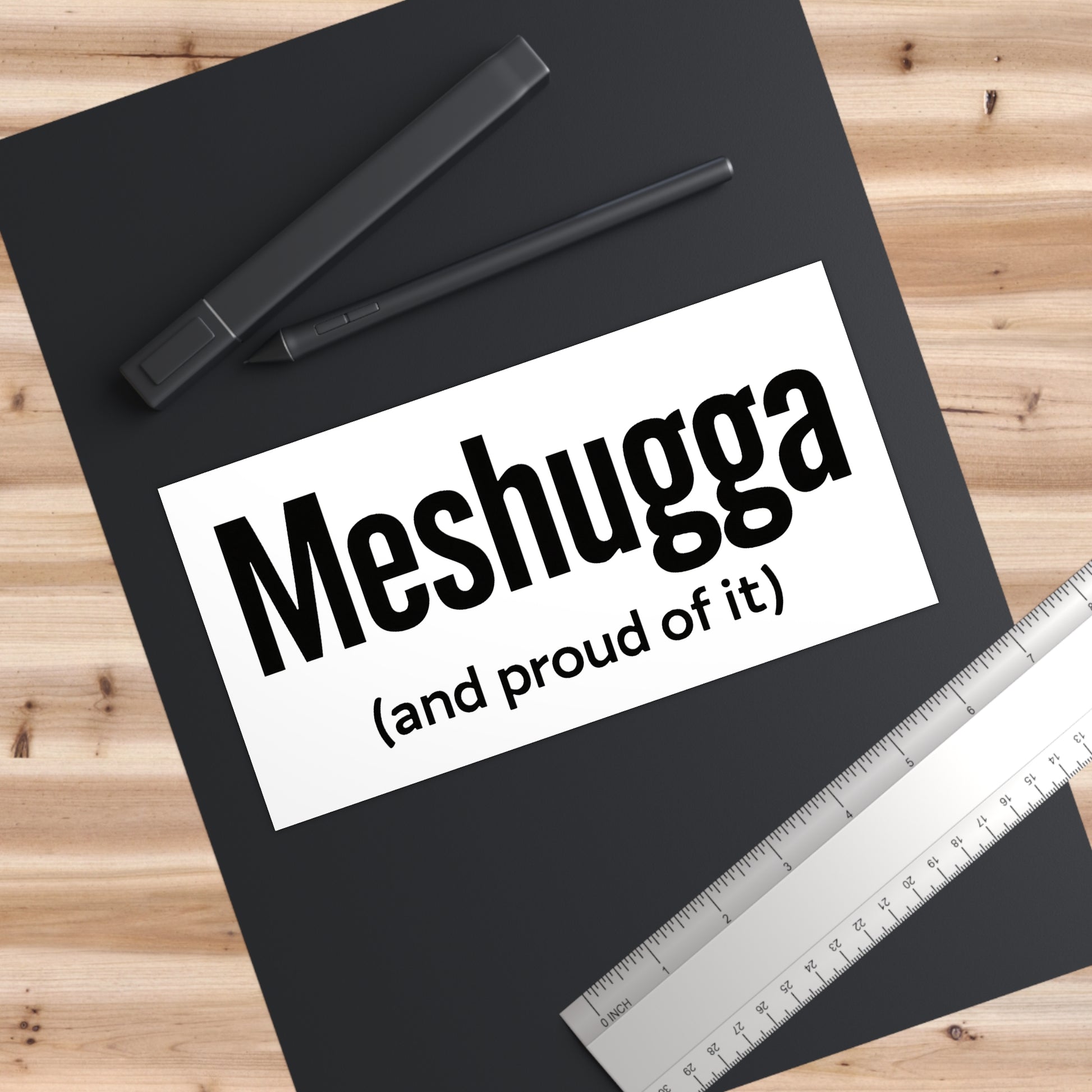 Black notebook with 'Meshugga (and proud of it)' text on a wooden surface with pens and a ruler.