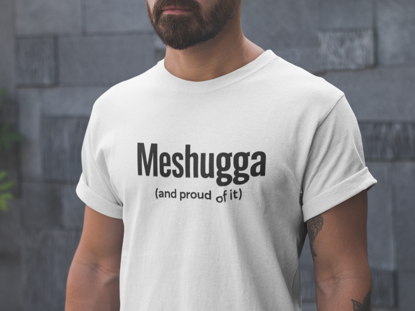 Man wearing a white t-shirt with 'Meshugga (and proud of it)' text against a stone wall.