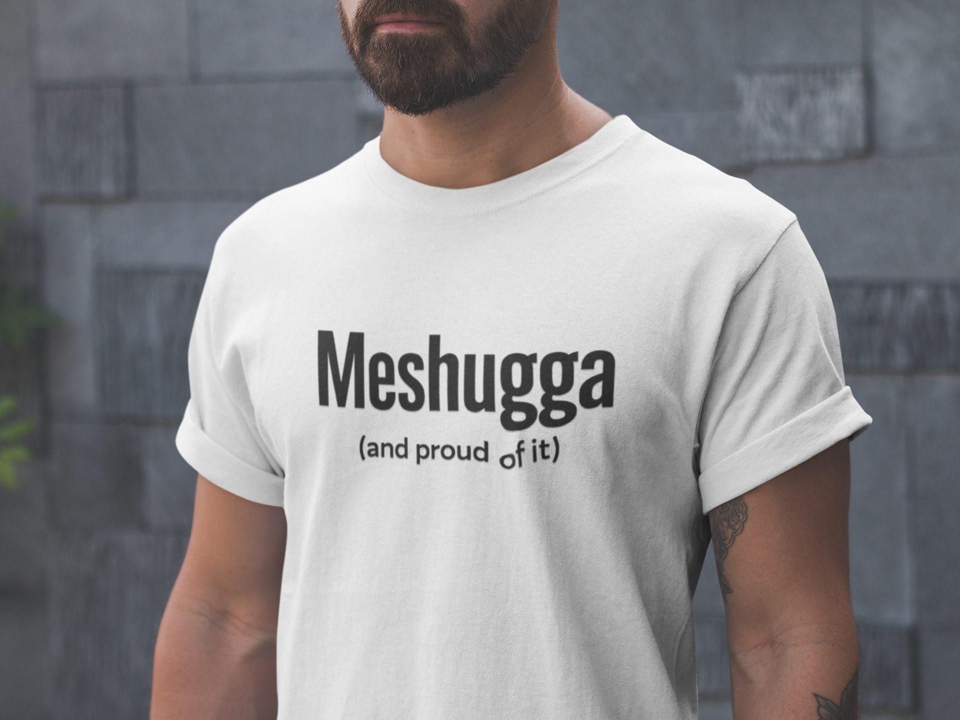 Man wearing a white t-shirt with 'Meshugga (and proud of it)' text against a stone wall.