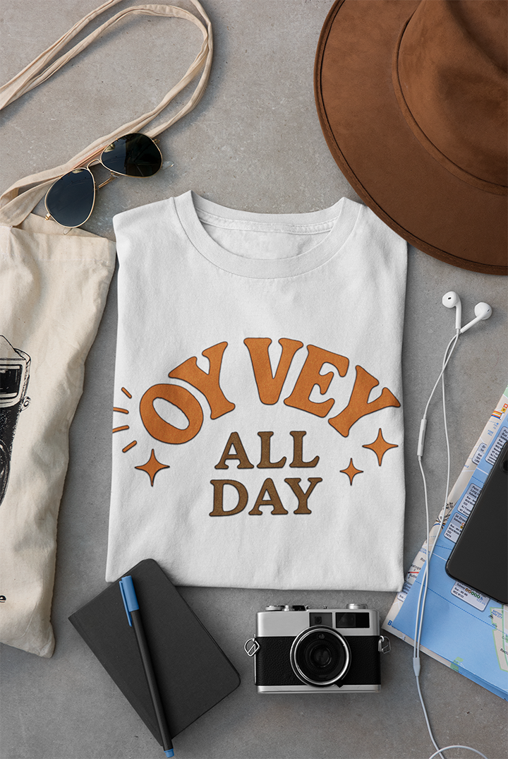 White t-shirt with 'Oy Vey All Day' text on a gray surface with accessories.