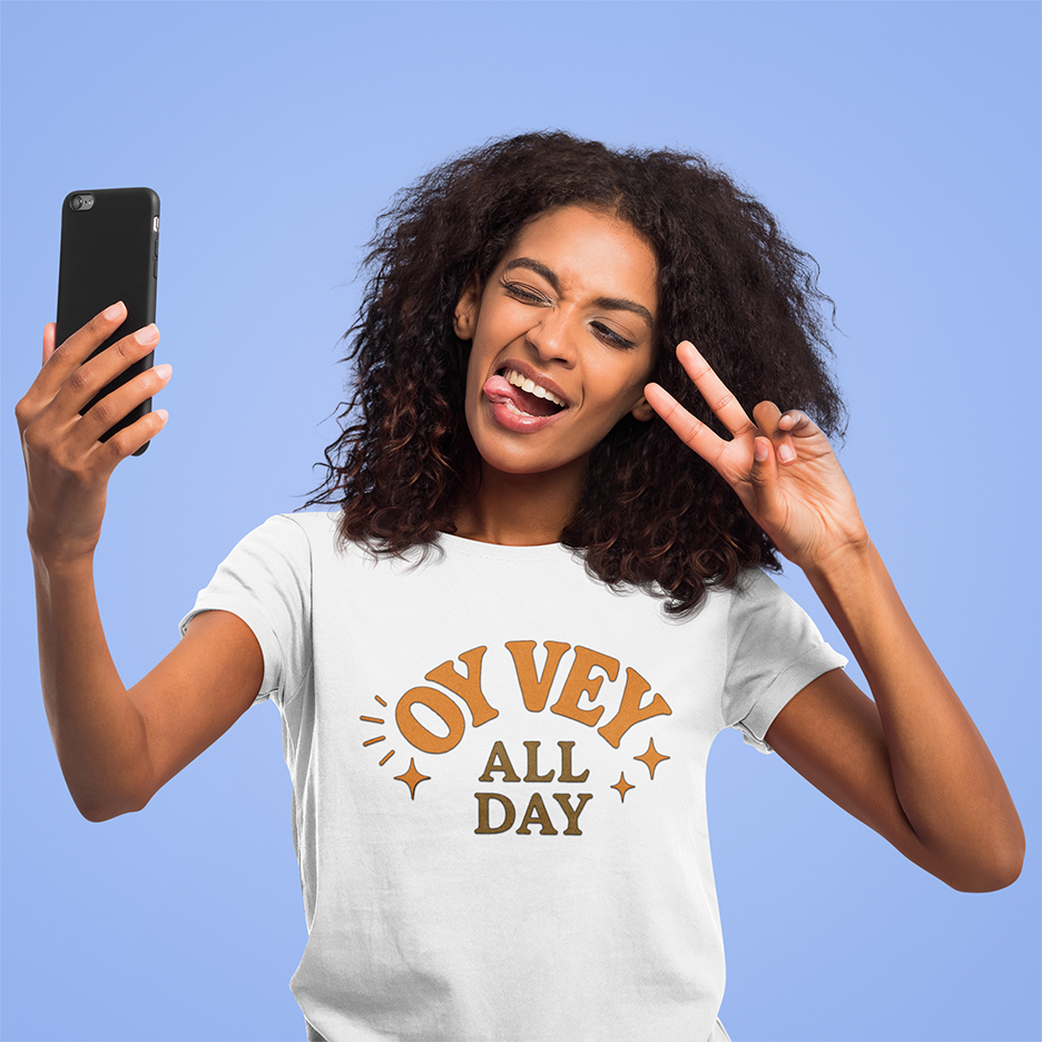 Woman wearing a 'Oy Vey All Day' t-shirt, making a peace sign and holding a phone against a blue background