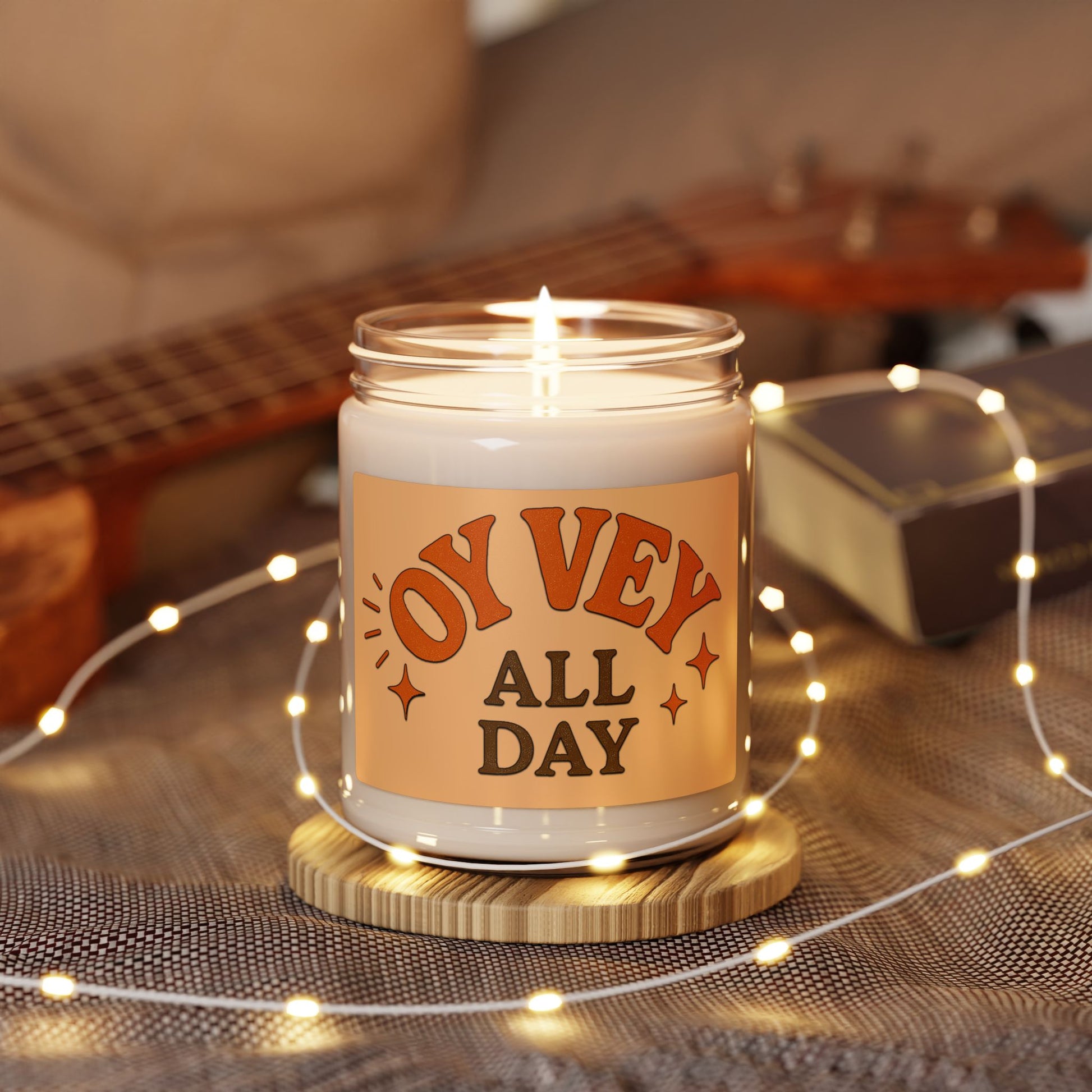 Candle in a jar with 'Oy Vey All Day' label on a wooden coaster with fairy lights, guitar in the background