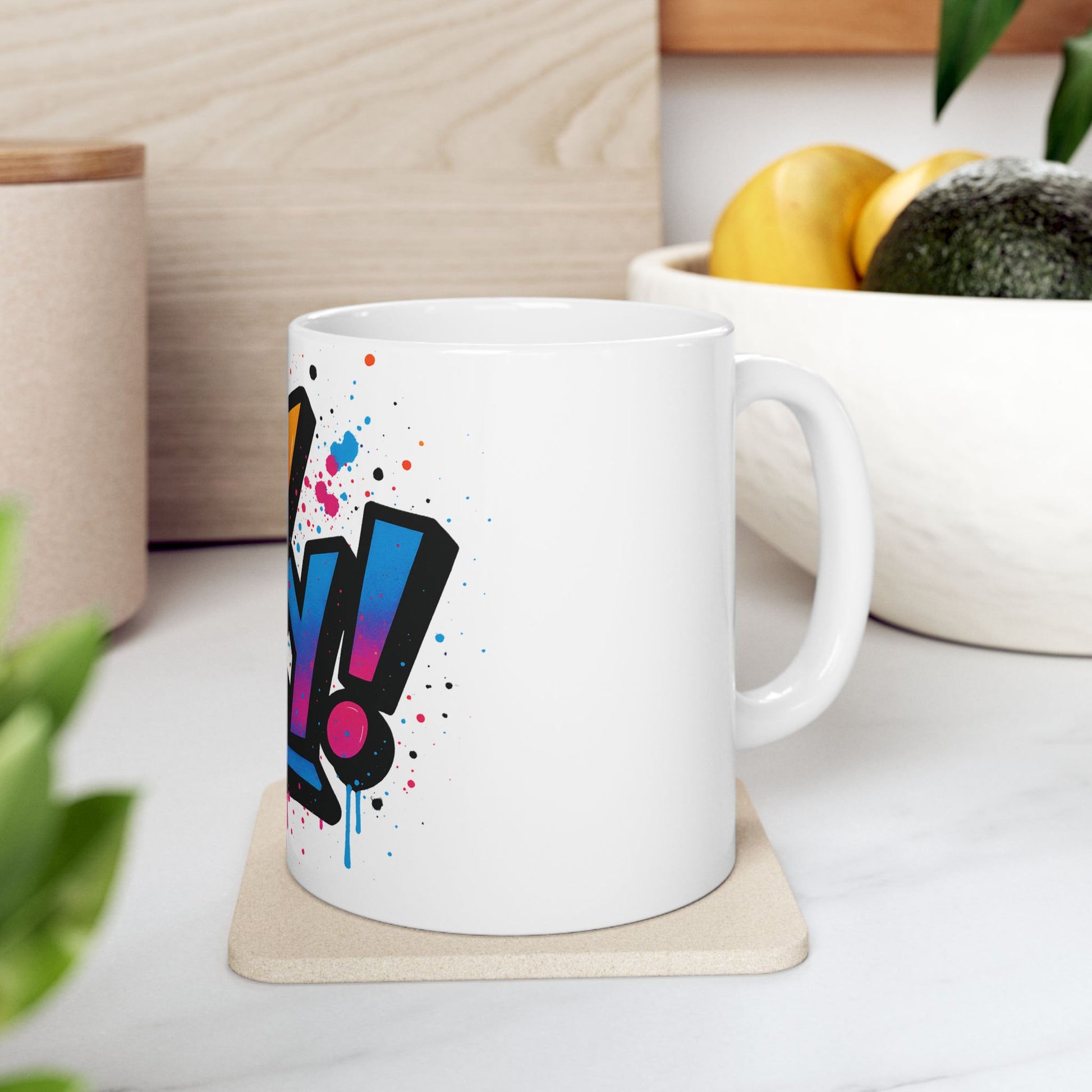 White mug with colorful 'oy vey' design on a coaster, placed on a surface with a bowl of fruit in the background.