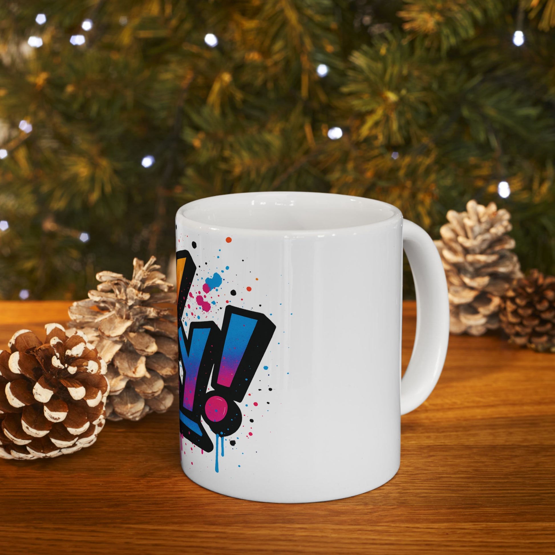 White mug with colorful 'oy vey' design on a wooden surface with pine cones and a Christmas tree in the background