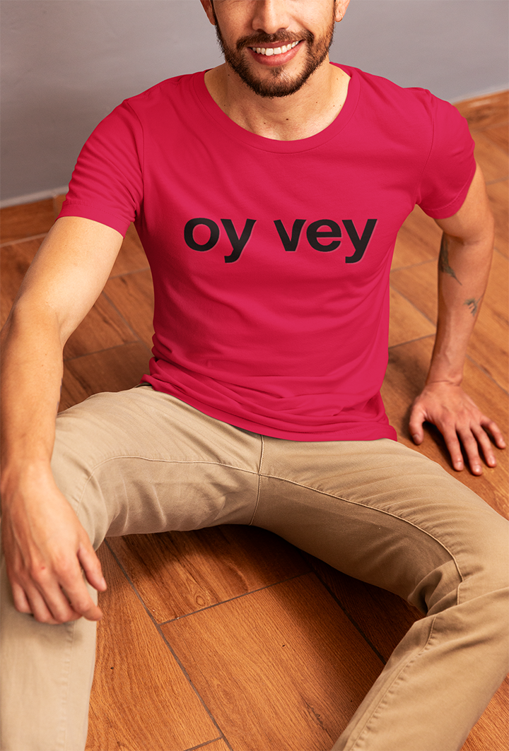 Man wearing a red t-shirt with 'oy vey' text sitting on a wooden floor.