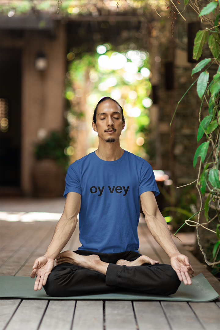 Man in a blue t-shirt with 'oy vey' text, sitting in a meditative pose on a mat outdoors.