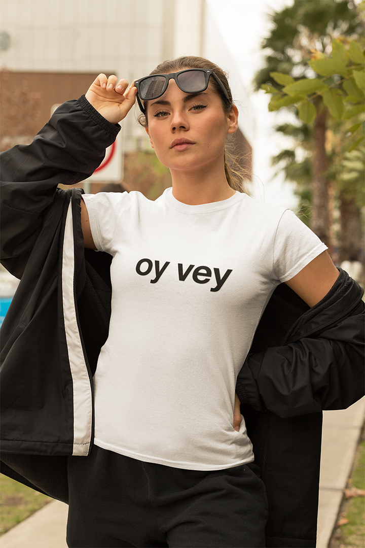 Person wearing a white t-shirt with 'oy vey' text, holding sunglasses, outdoors.