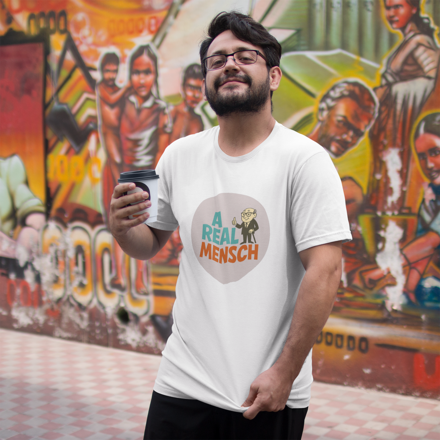 Man wearing a 'A Real Mensch' t-shirt holding a coffee cup in front of a colorful mural.