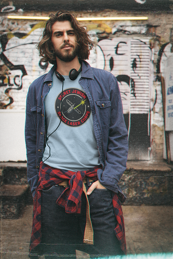 Man wearing a denim jacket and graphic t-shirt with a checkered scarf, standing in front of graffiti.