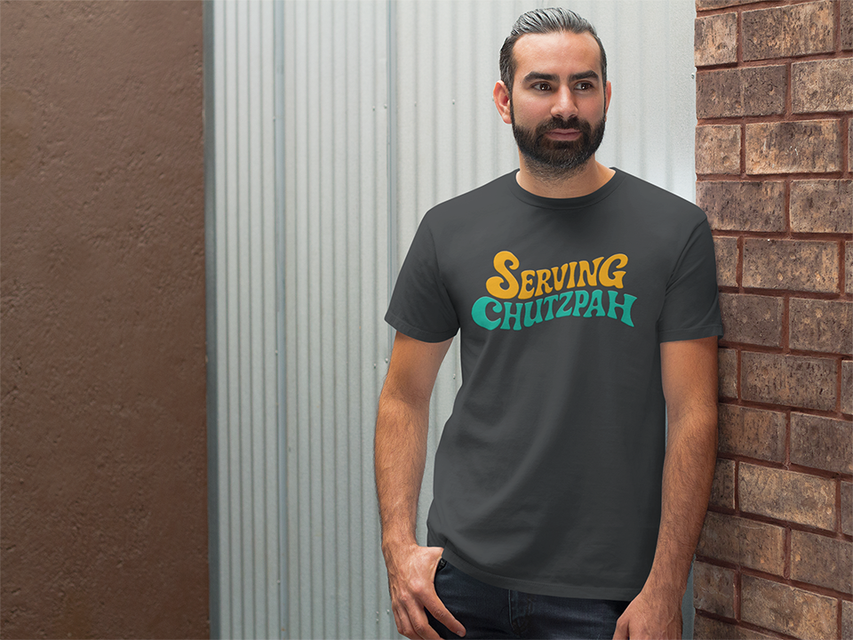 Man wearing a t-shirt with 'Serving Chutzpah' text standing against a brick wall.