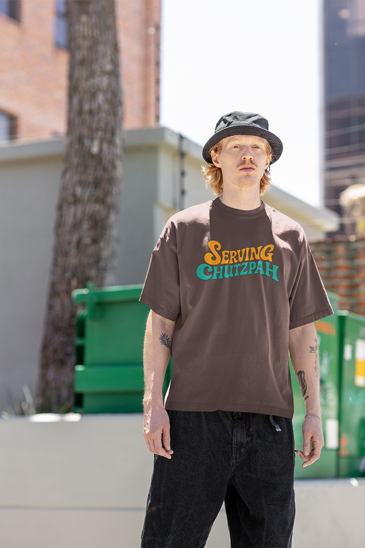 Person wearing a brown 'serving chutzpah' t-shirt with text, standing outdoors with buildings in the background