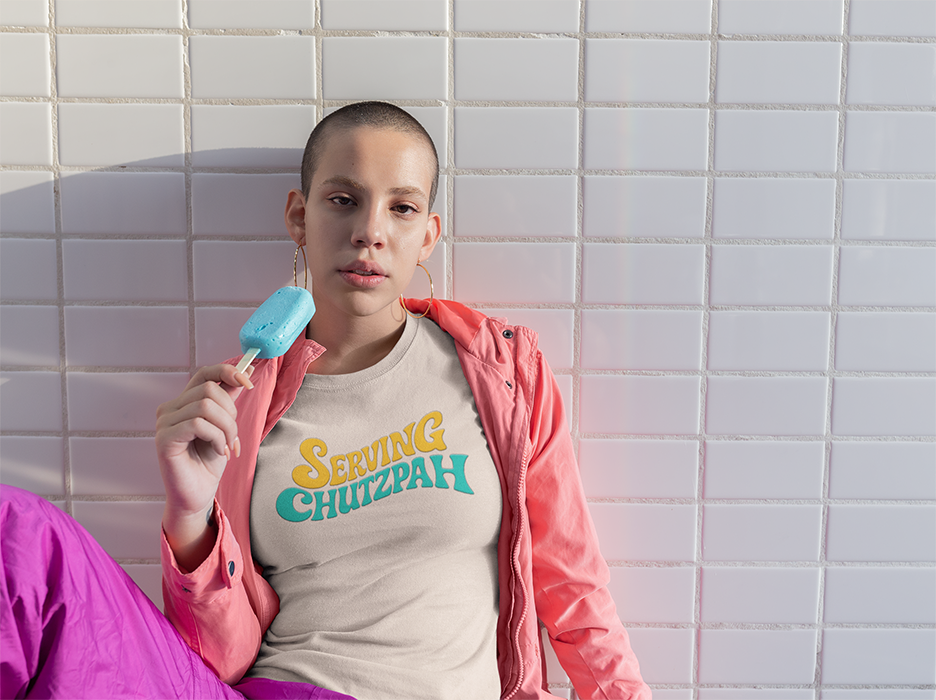 Person wearing a shirt with 'Serving Chutzpah' text, holding a popsicle against a tiled wall.
